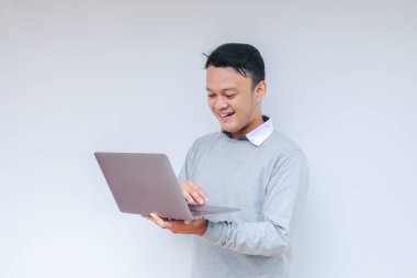 Genç Asyalı adam ayakta durup dizüstü bilgisayarla çalışırken gülümsüyor. Gri gömlekli Endonezyalı adam.