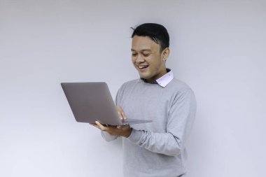 Genç Asyalı adam ayakta durup dizüstü bilgisayarla çalışırken gülümsüyor. Gri gömlekli Endonezyalı adam.