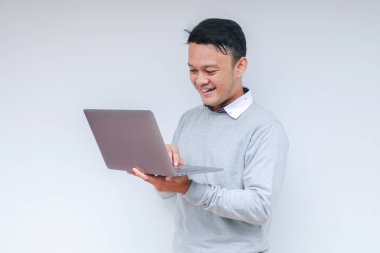 Genç Asyalı adam ayakta durup dizüstü bilgisayarla çalışırken gülümsüyor. Gri gömlekli Endonezyalı adam.