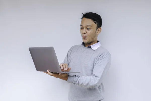 Wow face of Young Asian man shocked what he see in the laptop when working isolated grey background wearing grey shirt.