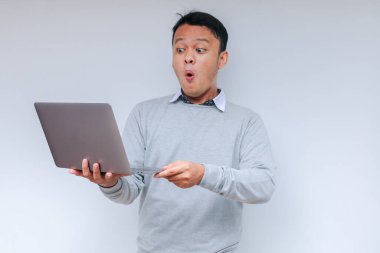 Genç Asyalı adamın yüzü, gri gömlekli, izole edilmiş gri arka planda çalışırken bilgisayarında gördüklerini şok etti..