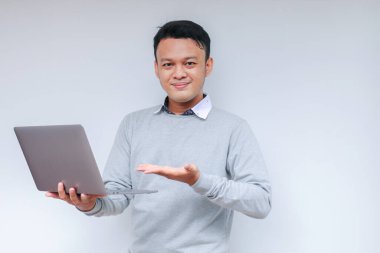 Genç Asyalı adam ayakta durup dizüstü bilgisayarla çalışırken gülümsüyor. Gri gömlekli Endonezyalı adam.