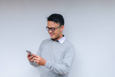 Zeki Asyalı genç adam stüdyo arka planında akıllı telefon kullanırken mutlu ve gülümsüyor.