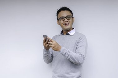 Zeki Asyalı genç adam stüdyo arka planında akıllı telefon kullanırken mutlu ve gülümsüyor.