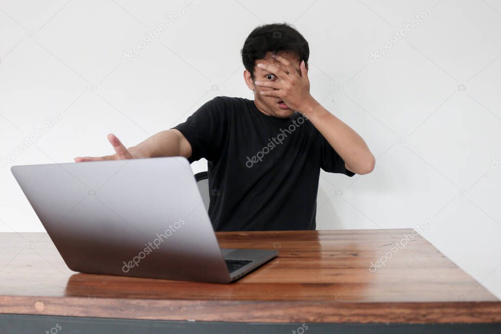 Young asian man hiding his face with hand because shocked and embarrassed by some porn videos or another forbidden thing he saw on the internet using a laptop