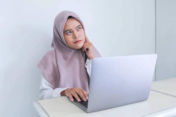 身穿头巾的年轻的亚洲伊斯兰女性 带着思考和仰视模仿空间 把一个手指放在脸颊上 具有灰色背景的成功的印度尼西亚伊斯兰妇女 — 图库照片