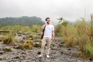 Genç Asyalı sırt çantalı adam mutlu ve dağda seyahat ederken akıllı telefona baktığında gülümsüyor.