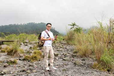 Genç Asyalı sırt çantalı adam mutlu ve dağda seyahat ederken akıllı telefona baktığında gülümsüyor.