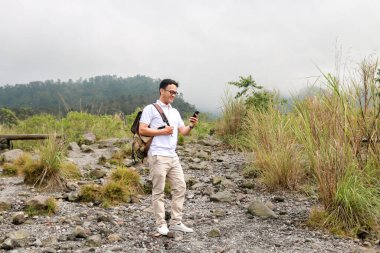 Genç Asyalı sırt çantalı adam mutlu ve dağda seyahat ederken akıllı telefona baktığında gülümsüyor.