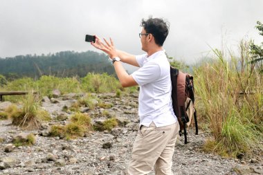 Genç Asyalı sırt çantalı adam dağda seyahat ederken video veya video çekmekten memnun.