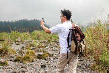 Genç Asyalı sırt çantalı adam dağda seyahat ederken video veya video çekmekten memnun.