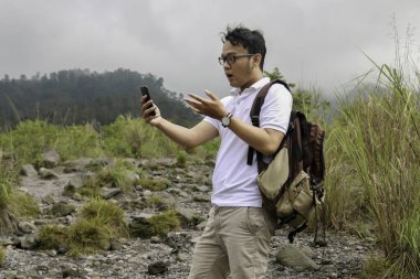 Genç Asyalı sırt çantalı adam dağda seyahat ederken akıllı telefona baktığında şok olur.