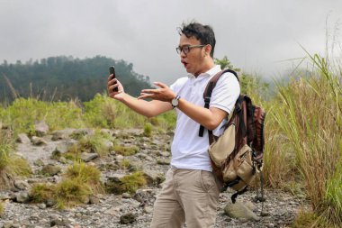 Genç Asyalı sırt çantalı adam dağda seyahat ederken akıllı telefona baktığında şok olur.