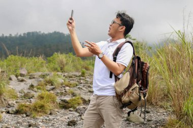 Genç Asyalı sırt çantalı adam dağda seyahat ederken akıllı telefona baktığında şok olur.