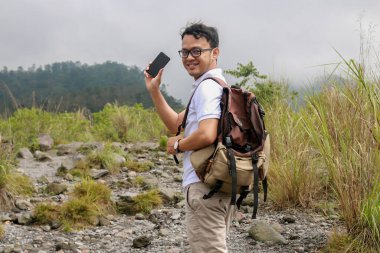 Genç Asyalı sırt çantalı adam mutlu ve dağda seyahat ederken akıllı telefon gösterdiğinde gülümsüyor.