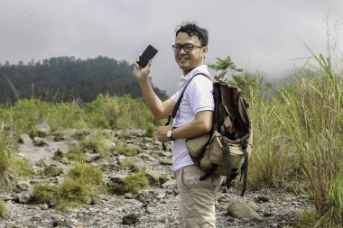 Genç Asyalı sırt çantalı adam mutlu ve dağda seyahat ederken akıllı telefon gösterdiğinde gülümsüyor.