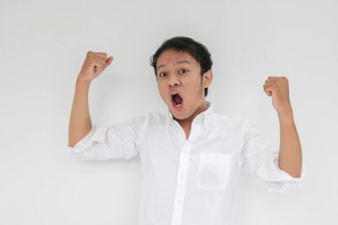 Mutlu ve gülümseyen Asyalı genç adam başarı ve başarıyı kutlamak için kolunu kaldırıyor. Beyaz gömlek giyen Endonezyalı adam