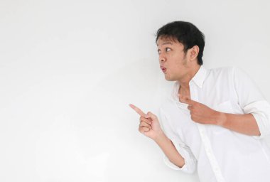 Genç Asyalı adamın beyaz gömleği şaşırdı ve haykırıyor. Vay canına! Parmağıyla gri arka planı işaret ediyor.