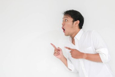 Genç Asyalı adamın beyaz gömleği şaşırdı ve haykırıyor. Vay canına! Parmağıyla gri arka planı işaret ediyor.