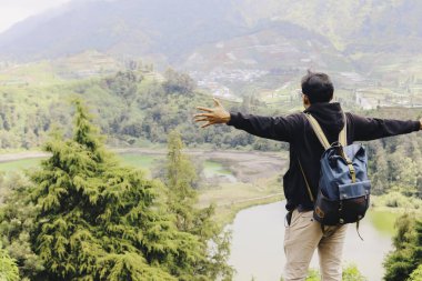 Genç adam bir uçurumun kenarında duruyor ve Dieng Endonezya 'daki dağ ve göl manzarasının tadını çıkarıyor..