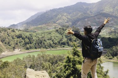 Genç adam bir uçurumun kenarında duruyor ve Dieng Endonezya 'daki dağ ve göl manzarasının tadını çıkarıyor..
