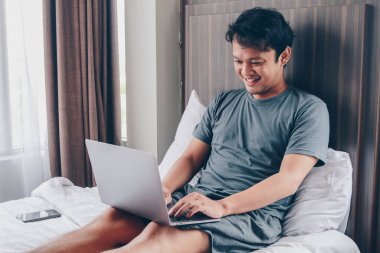 Mutlu Asyalı adam laptopuyla yatağında çalışıyor. Serbest çalışan başarılı yaşam tarzı kavramı.