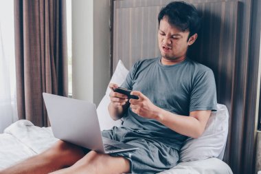 Genç Asyalı adam dizüstü bilgisayarda oyun oynarken üzgün ve sinirli ve yatakta telefonla konuşuyor.