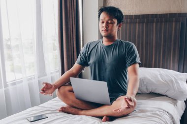 Evden çalışıyorum. Genç Asyalı bir adam yatakta oturuyor. Ev işyerinde. Mola, iş zamanı rahatlama..