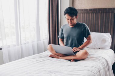 Ciddi Asyalı adam laptopuyla yatağında çalışıyor. Serbest çalışan başarılı yaşam tarzı kavramı.