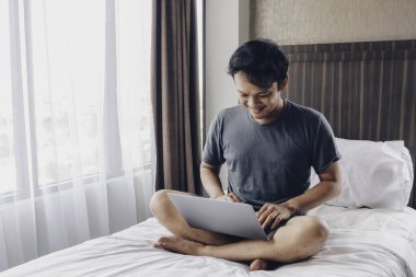 Mutlu Asyalı adam laptopuyla yatağında çalışıyor. Serbest çalışan başarılı yaşam tarzı kavramı.