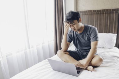 Yatak odasındaki dizüstü bilgisayarı kullanırken yorgun ve yorgun hisseden Asyalı bir adam. Sıkı çalışma konsepti.