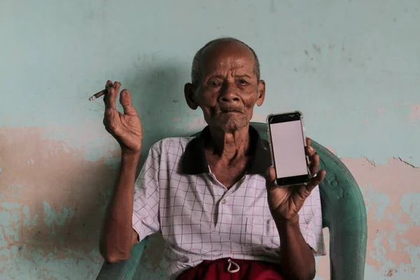 A portrait of Indonesian old grandpa showing white screen smartphone.