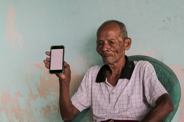 A portrait of Indonesian old grandpa showing white screen smartphone.