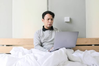 Genç Asyalı adam dizüstü bilgisayarda oyun oynarken üzgün ve sinirli ve yatakta telefonla konuşuyor.