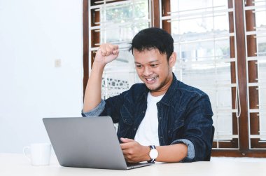 Mutlu ve gülümseyen Asyalı genç adam laptopta çalışırken başarıyı ya da başarıyı kutlamak için kolunu kaldırıyor. Endonezyalı adam mavi gömlek giyiyor..