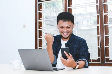 Mutlu ve gülümseyen Asyalı genç adam laptopta çalışırken başarıyı ya da başarıyı kutlamak için kolunu kaldırıyor. Endonezyalı adam mavi gömlek giyiyor..
