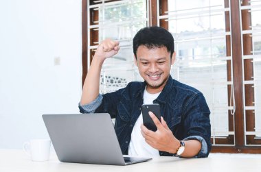 Mutlu ve gülümseyen Asyalı genç adam laptopta çalışırken başarıyı ya da başarıyı kutlamak için kolunu kaldırıyor. Endonezyalı adam mavi gömlek giyiyor..