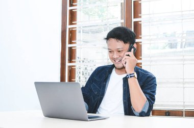 Genç Asyalı adam telefon ve dizüstü bilgisayarla çalışırken mutlu ve gülümsüyor..