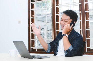 Genç Asyalı adam mutlu ve akıllı telefondan arandığında laptopta gördüklerine gülümsüyor.