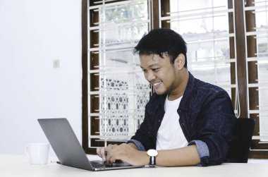 Genç Asyalı adam kendini mutlu hissediyor ve laptopta çalışırken gülümsüyor. Endonezyalı adam mavi gömlek giyiyor..