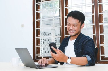 Genç Asyalı adam telefon ve dizüstü bilgisayarla çalışırken mutlu ve gülümsüyor..