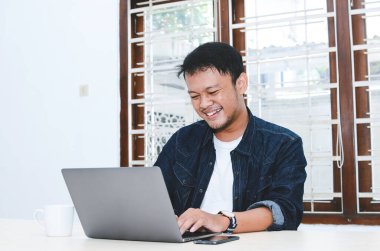 Genç Asyalı adam kendini mutlu hissediyor ve laptopta çalışırken gülümsüyor. Endonezyalı adam mavi gömlek giyiyor..