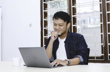 Genç Asyalı adam kendini mutlu hissediyor ve laptopta çalışırken gülümsüyor. Endonezyalı adam mavi gömlek giyiyor..