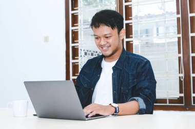 Genç Asyalı adam kendini mutlu hissediyor ve laptopta çalışırken gülümsüyor. Endonezyalı adam mavi gömlek giyiyor..