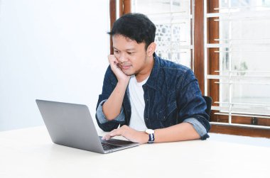 Genç Asyalı adam kendini mutlu hissediyor ve laptopta çalışırken gülümsüyor. Endonezyalı adam mavi gömlek giyiyor..