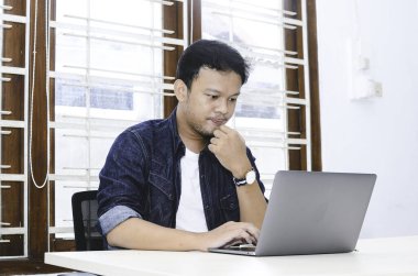 Genç Asyalı adam masada dizüstü bilgisayarla çalışırken ciddi hissediyor.