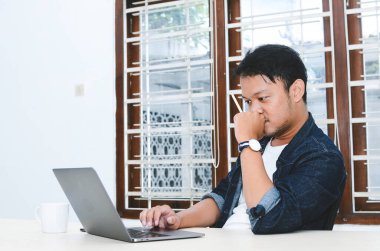 Genç Asyalı adam masada dizüstü bilgisayarla çalışırken ciddi hissediyor.