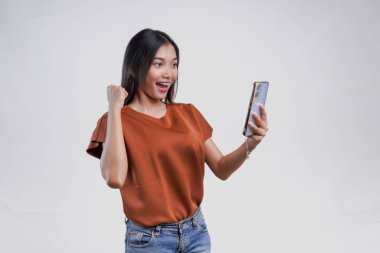 Kahverengi tişörtlü neşeli genç Asyalı kadın mutlu bir ifadeyle akıllı telefon tutuyor, beyaz arka planla izole edilmiş..