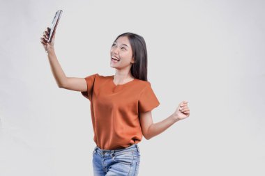 Kahverengi tişörtlü neşeli genç Asyalı kadın mutlu bir ifadeyle akıllı telefon tutuyor, beyaz arka planla izole edilmiş..