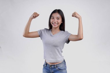 Gri gömlekli kendine güvenen Asyalı kadın gururla gülümserken kollarını ve kaslarını kaldırarak güçlü bir hareket sergiliyor..